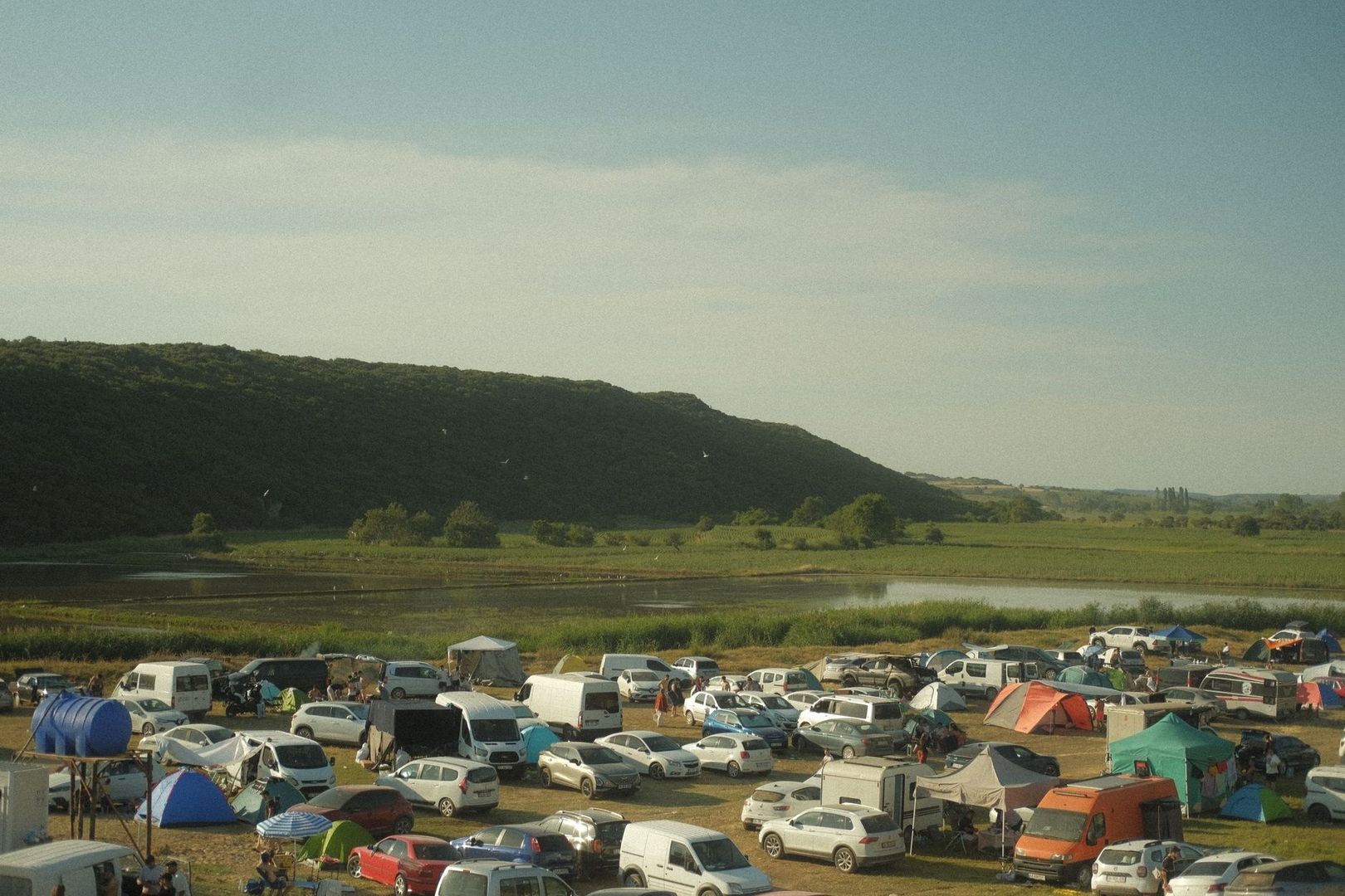 Crowded lakeside campsite with tents, vehicles, fields, and wooded hills